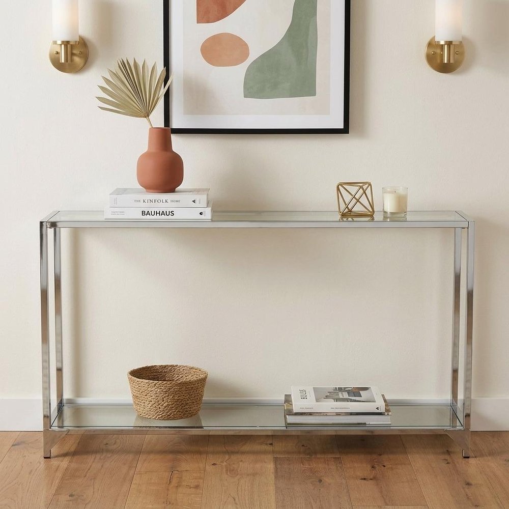 56" Clear and Silver Glass and Iron Frame Console Table With Shelf styled in a furnished interior with an off-white wall on a dark wood floor.