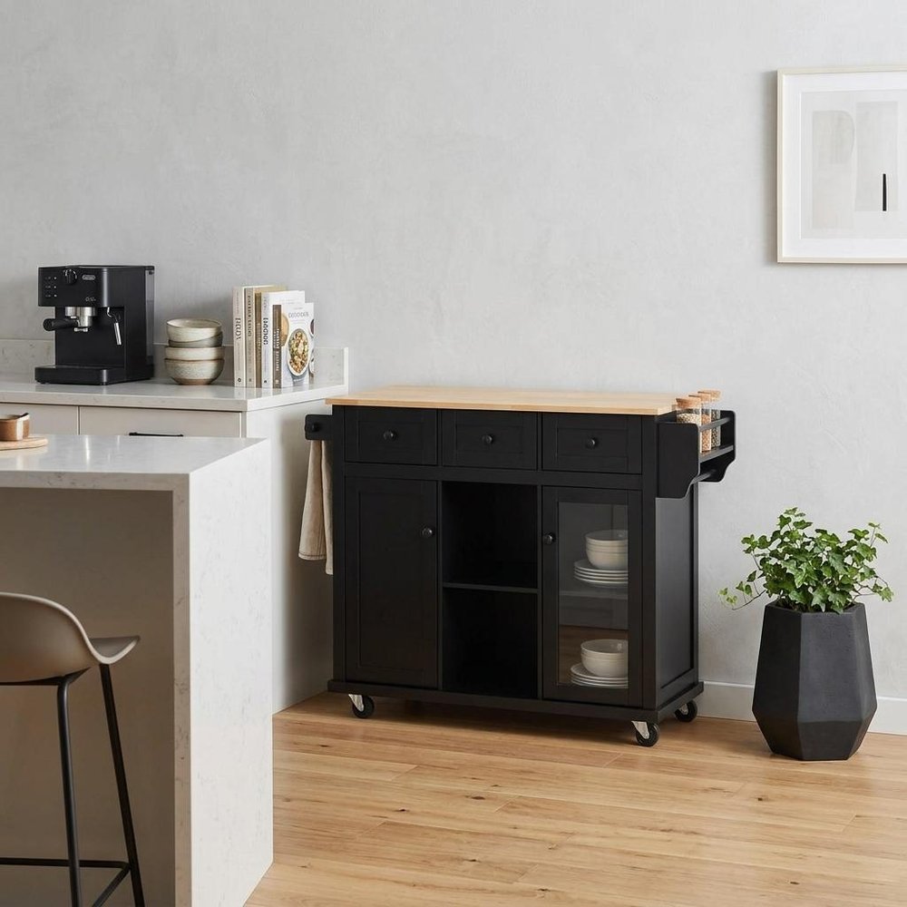 53" Brown and Black Wood Flat Door Rolling Kitchen Cart With Storage styled in a furnished interior with a light gray wall on a wood floor.